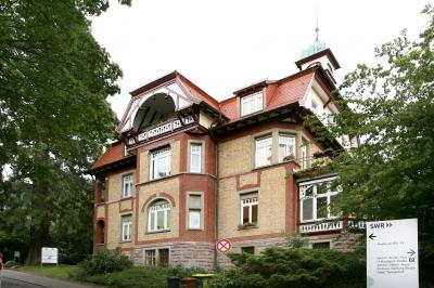 Ein großes, reich verziertes Backsteingebäude mit rotem Dach steht zwischen üppigen grünen Bäumen und ähnelt der komplizierten Präzision von Zahnersatz. Das Gebäude hat mehrere Stockwerke mit weiß gerahmten Fenstern und dekorativen Elementen. Im Vordergrund ist ein kleines Schild mit Richtungspfeilen zu sehen.