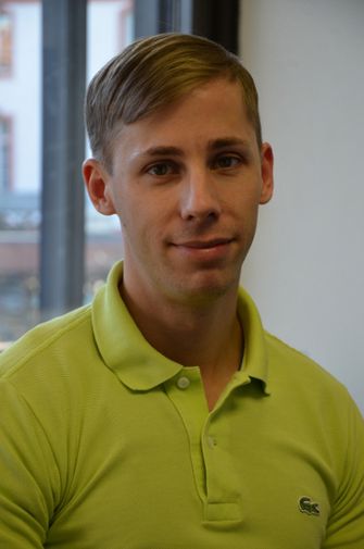 Eine Person in einem hellgrünen Poloshirt steht drinnen vor einem Fenster, von draußen sind Stadtgebäude zu sehen. Mit neutralem Gesichtsausdruck blickt sie in die Kamera und denkt vielleicht über ihren nächsten Zahnersatz-Termin nach.