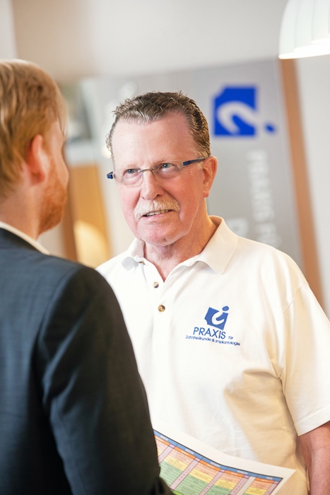 Ein Mann mit Brille und weißem Poloshirt mit Firmenlogo diskutiert mit einer anderen Person im Anzug über Zahnersatz. Im Hintergrund sind Teile eines Banners und eine verschwommene Inneneinrichtung zu sehen, die möglicherweise mit Lösungen für Zahnimplantate zu tun hat.