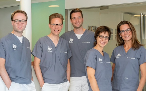 Fünf Personen in grauen Arztkitteln stehen zusammen im Innenbereich und lächeln in die Kamera. Es scheint sich um ein Team von auf Zahnersatz spezialisierten Gesundheitsexperten zu handeln, die in einer gut beleuchteten Umgebung arbeiten.